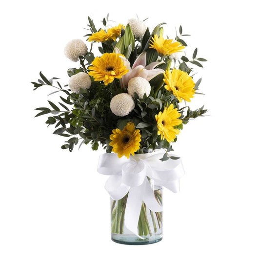 A bouquet of yellow gerberas, white pingpong flowers, and pink lilies arranged in a clear glass vase with a white ribbon.